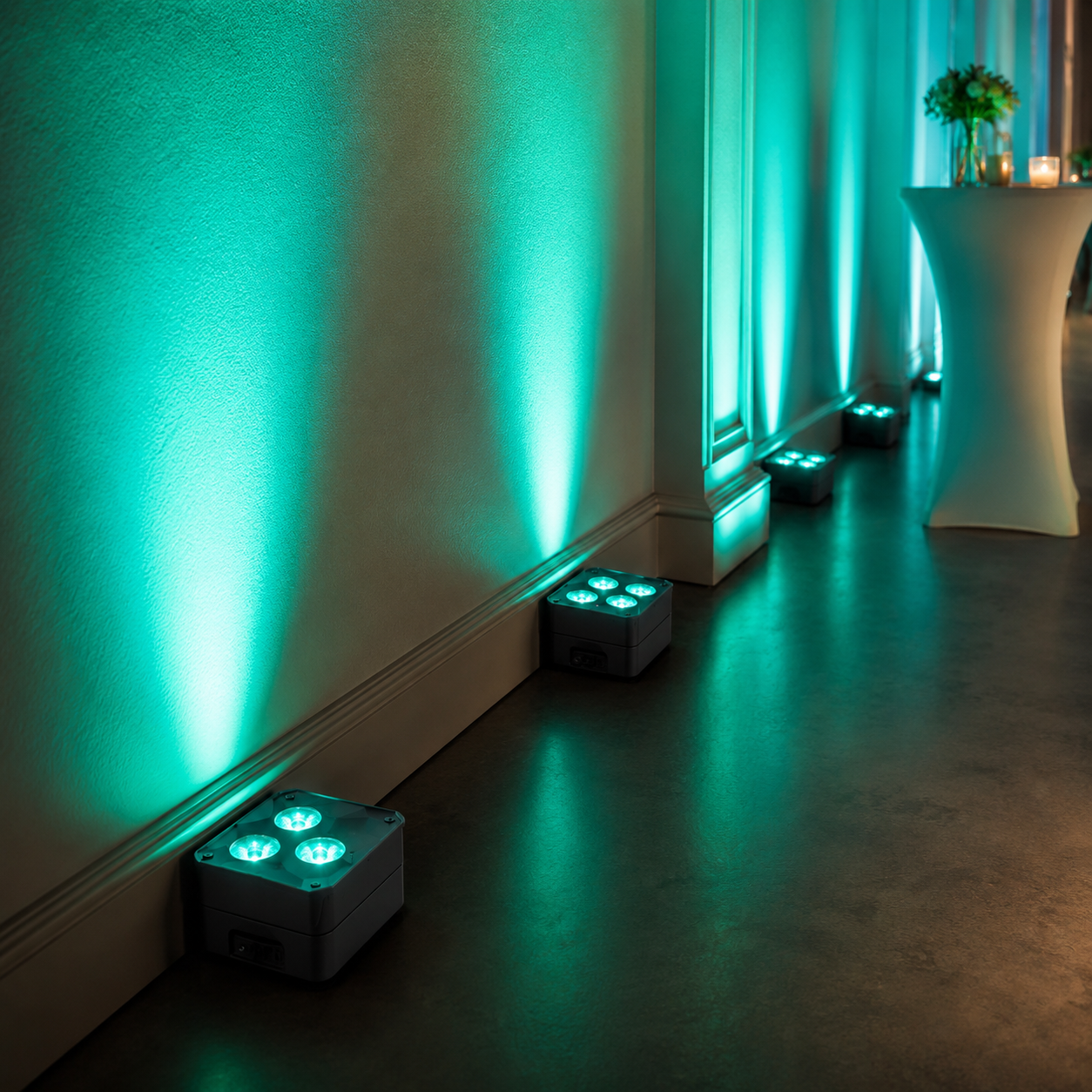 Teal uplighting along a wall with small square floor lights casting green glow and a tall cocktail table on the right with flowers and candles.
